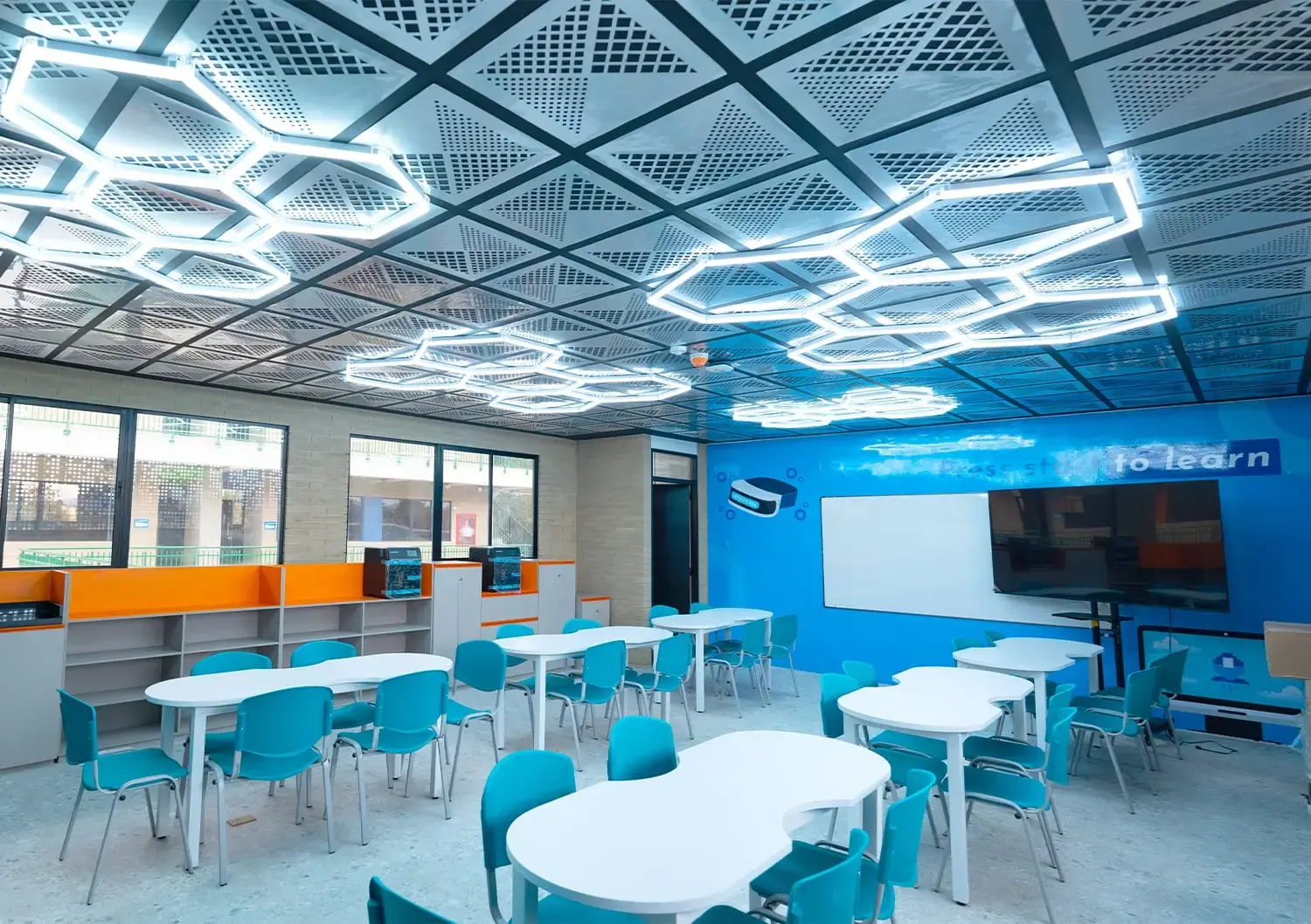 Aula interactiva con mesas de diseño modular, sillas celestes y un techo moderno con luces LED hexagonales.