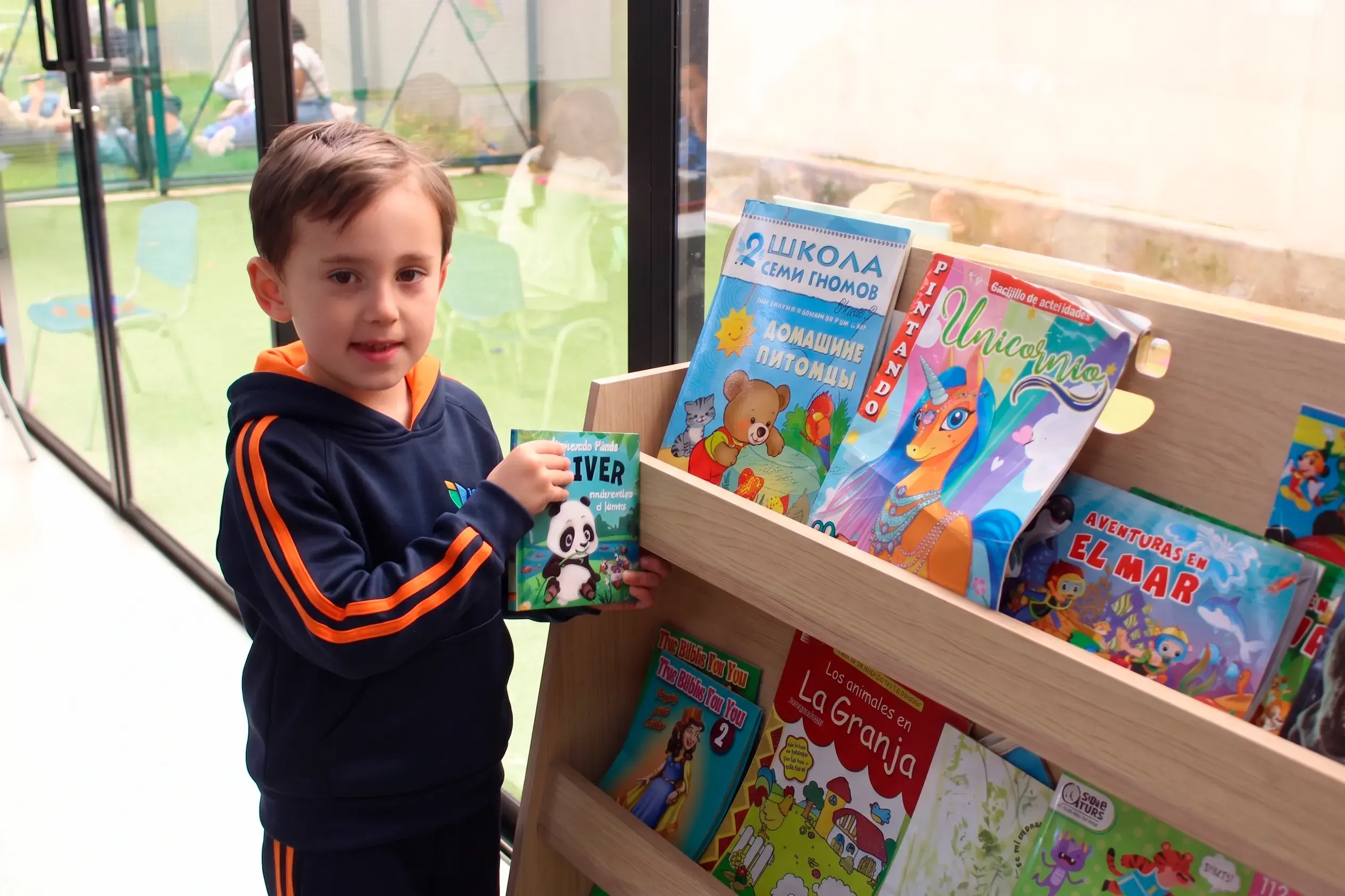 Un niño de primaria sonríe sosteniendo un libro infantil frente a un estante de madera con cuentos ilustrados en la biblioteca escolar del colegio.