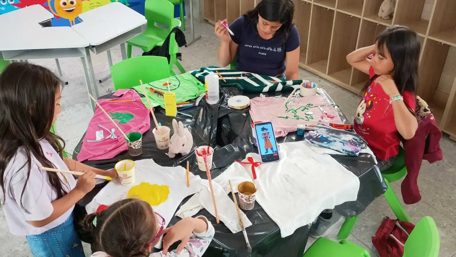 Grupo de niños pintando con pinceles sobre camisetas blancas extendidas en una mesa durante un taller de arte.