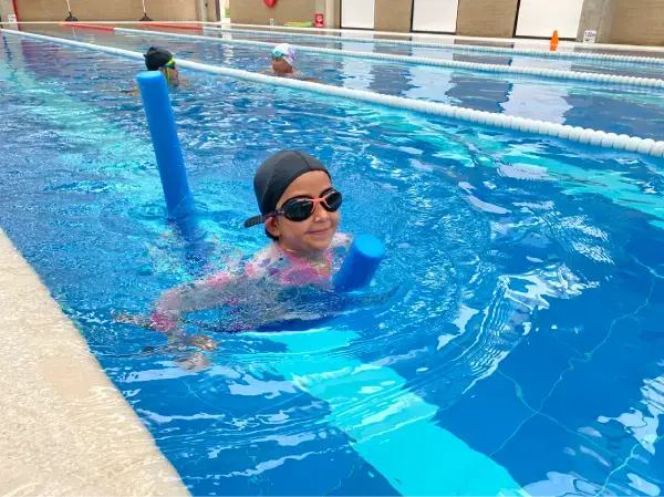 Niña sonriente practicando natación infantil con un flotador de tubo de espuma azul en una piscina con carriles.