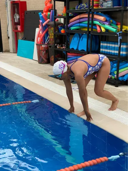 Niña pequeña con gorro rosa en el borde de la piscina preparándose para saltar o hacer un clavado al agua