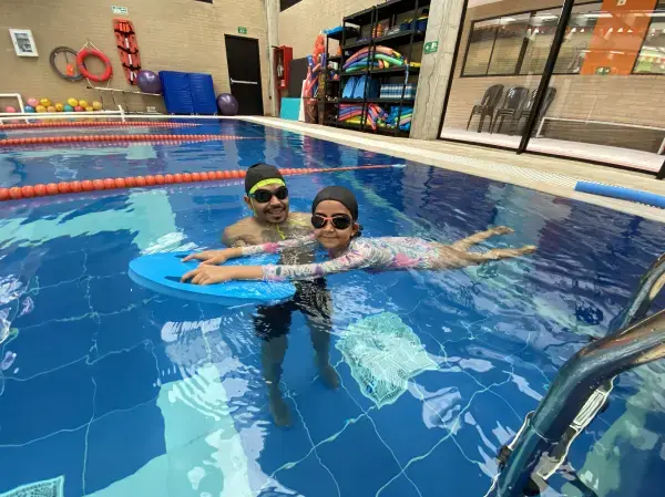 Profesor de natación guiando con cuidado a una niña que flota boca arriba en una piscina climatizada interior.