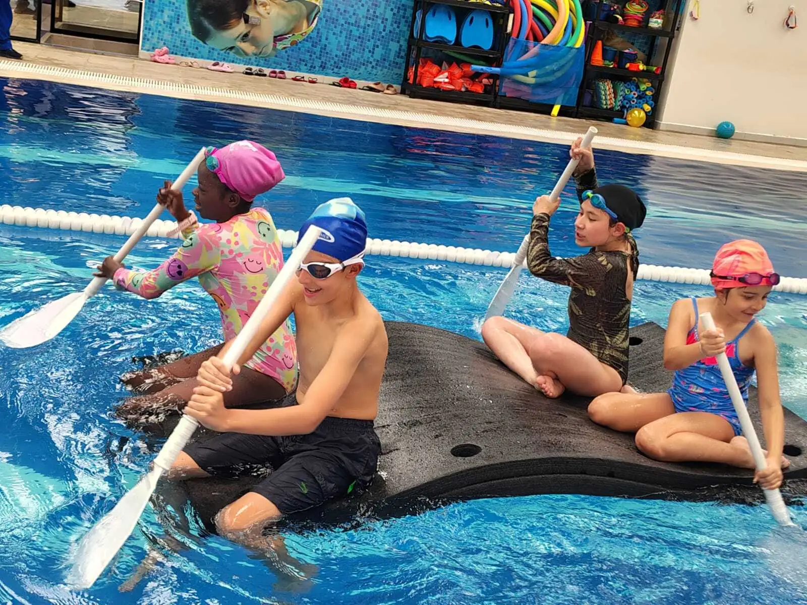 Cuatro niños pequeños divirtiéndose y remando sobre colchonetas flotantes en una clase lúdica de natación.