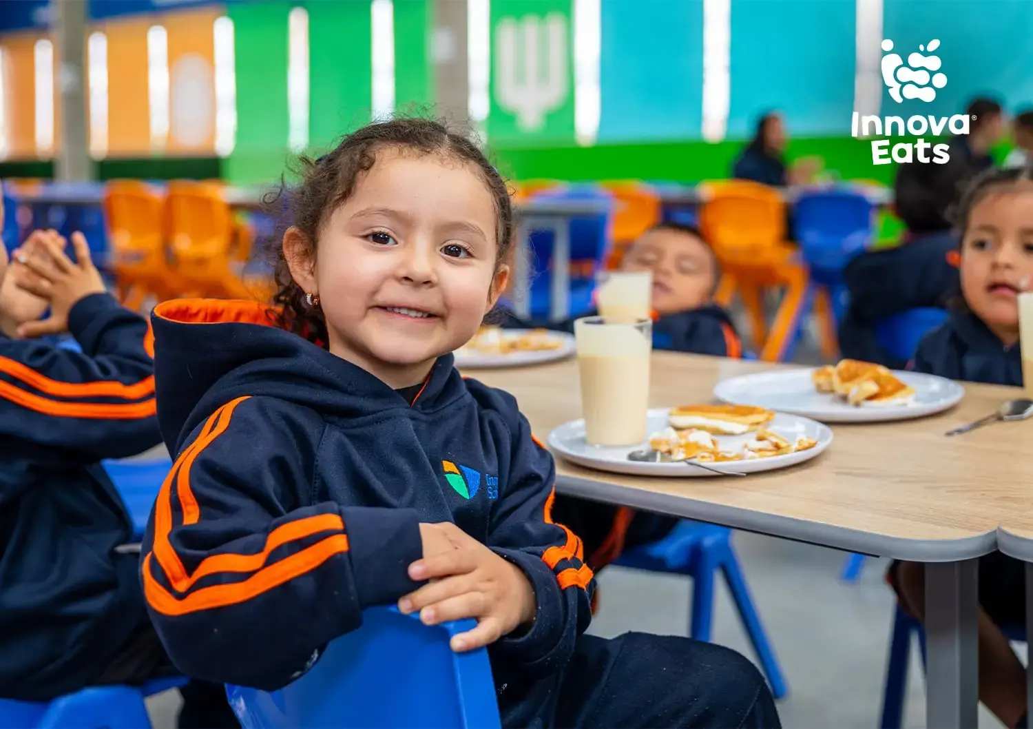 Niña sonriente sentada en el comedor escolar comiendo  de un plato equilibrado de Innova Eats.