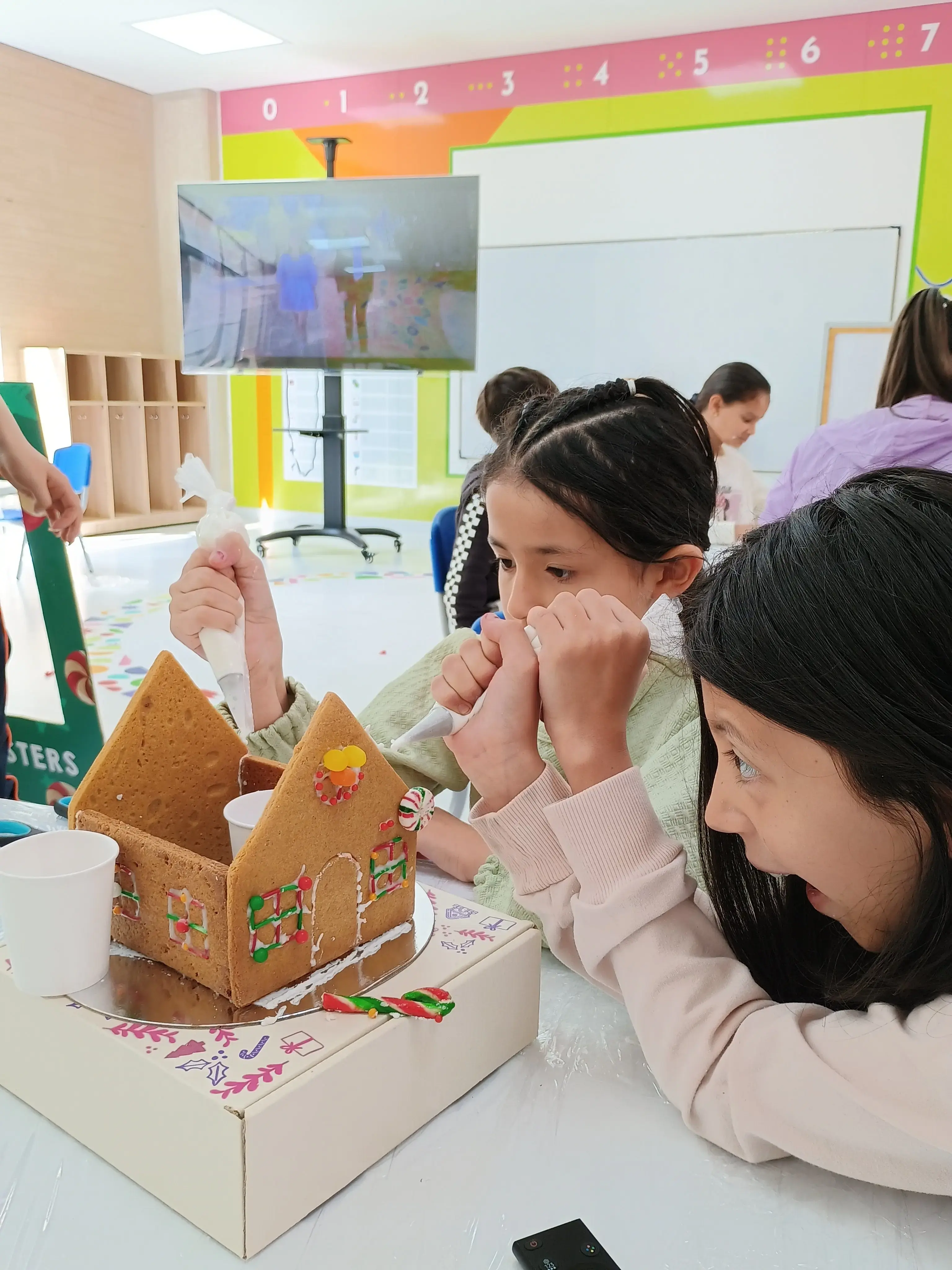 Dos niñas concentradas decorando una casa de galleta con dulces en un taller creativo de manualidades escolares.
